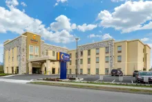  Gemini said The exterior of a multi-story Comfort Suites hotel with a tan and stone facade, featuring a large blue monument sign in the foreground under a bright, partly cloudy sky.