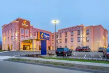 Wide-angle shot of the Comfort Suites in Burlington, Washington during twilight. The building features a modern design with tan siding and decorative stone pillars. A large blue and yellow Comfort Suites sign stands in the foreground, and the hotel parking lot is visible with several vehicles parked in front.