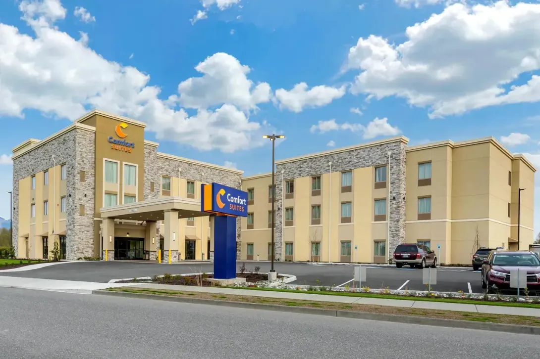  Gemini said The exterior of a multi-story Comfort Suites hotel with a tan and stone facade, featuring a large blue monument sign in the foreground under a bright, partly cloudy sky.