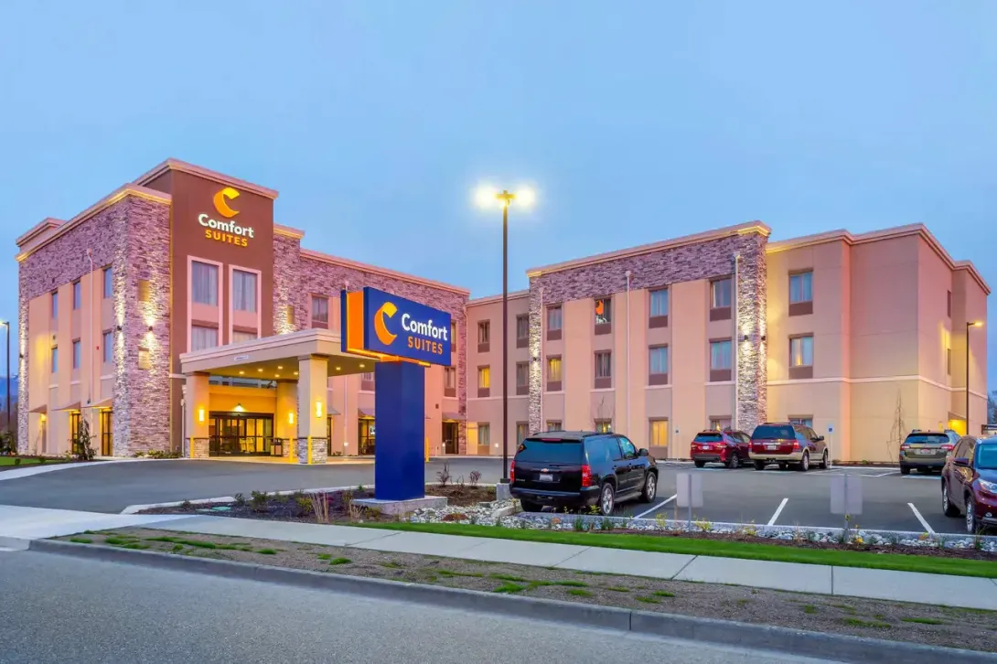 Wide-angle shot of the Comfort Suites in Burlington, Washington during twilight. The building features a modern design with tan siding and decorative stone pillars. A large blue and yellow Comfort Suites sign stands in the foreground, and the hotel parking lot is visible with several vehicles parked in front.